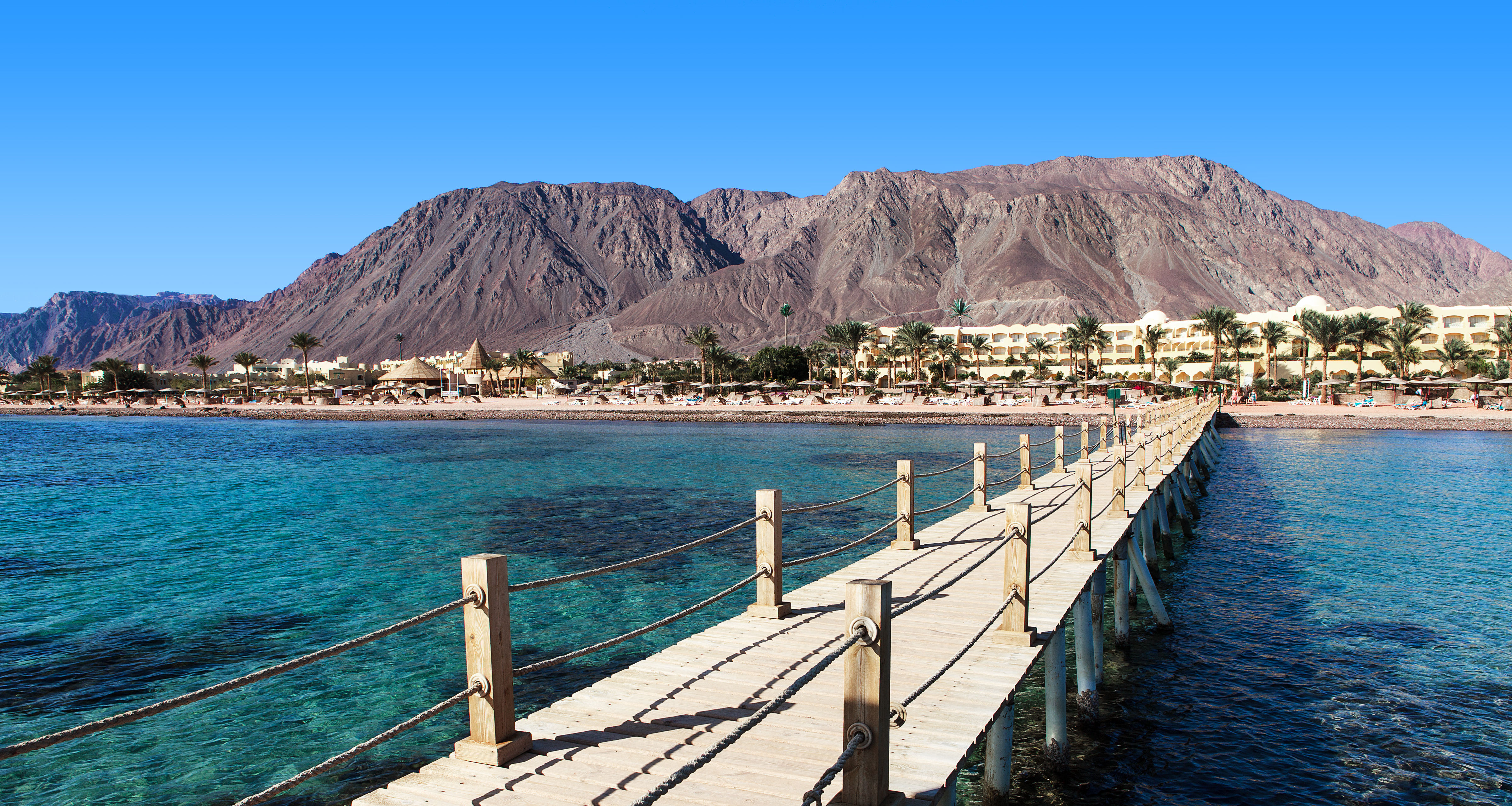 Pier over Red Sea reef with beach resort and mountain backdrop in Dahab Egypt.