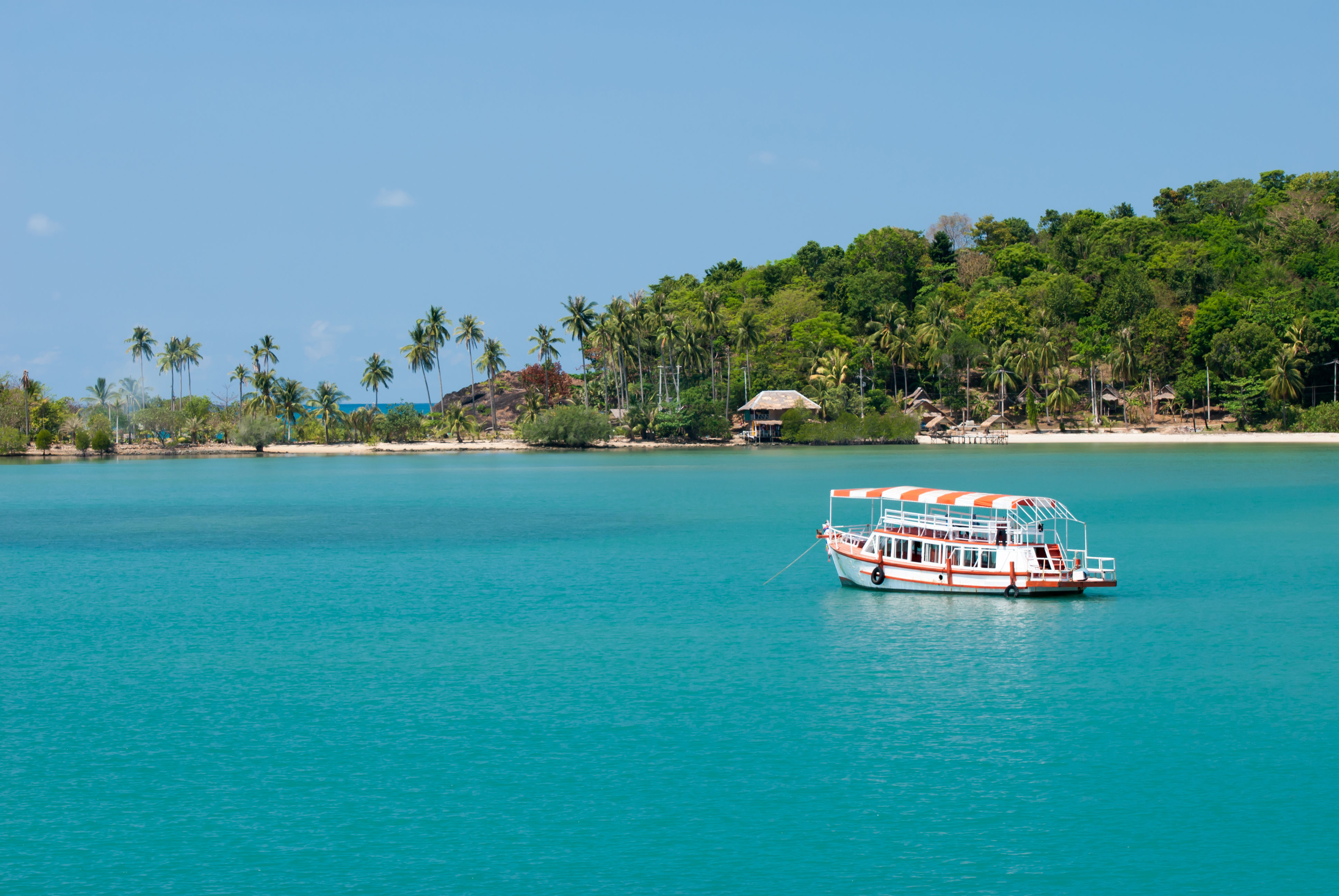 Tour boat sailing on turquoise waters near white sand beach and palm trees in Koh Chang Island.