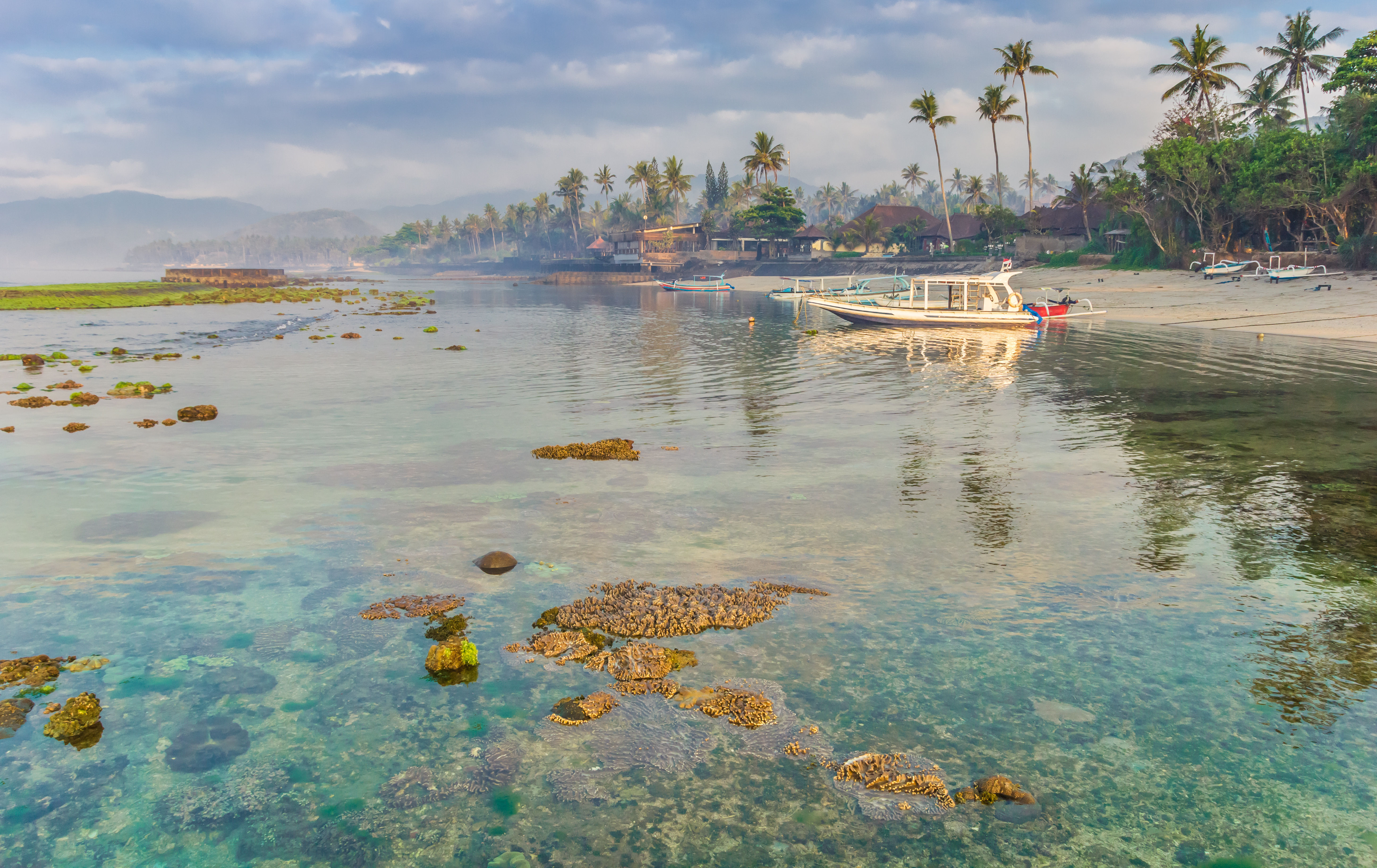 Shallow reef coral in crystal clear waters, moored boats and palm trees in Candi Dasa Beach.