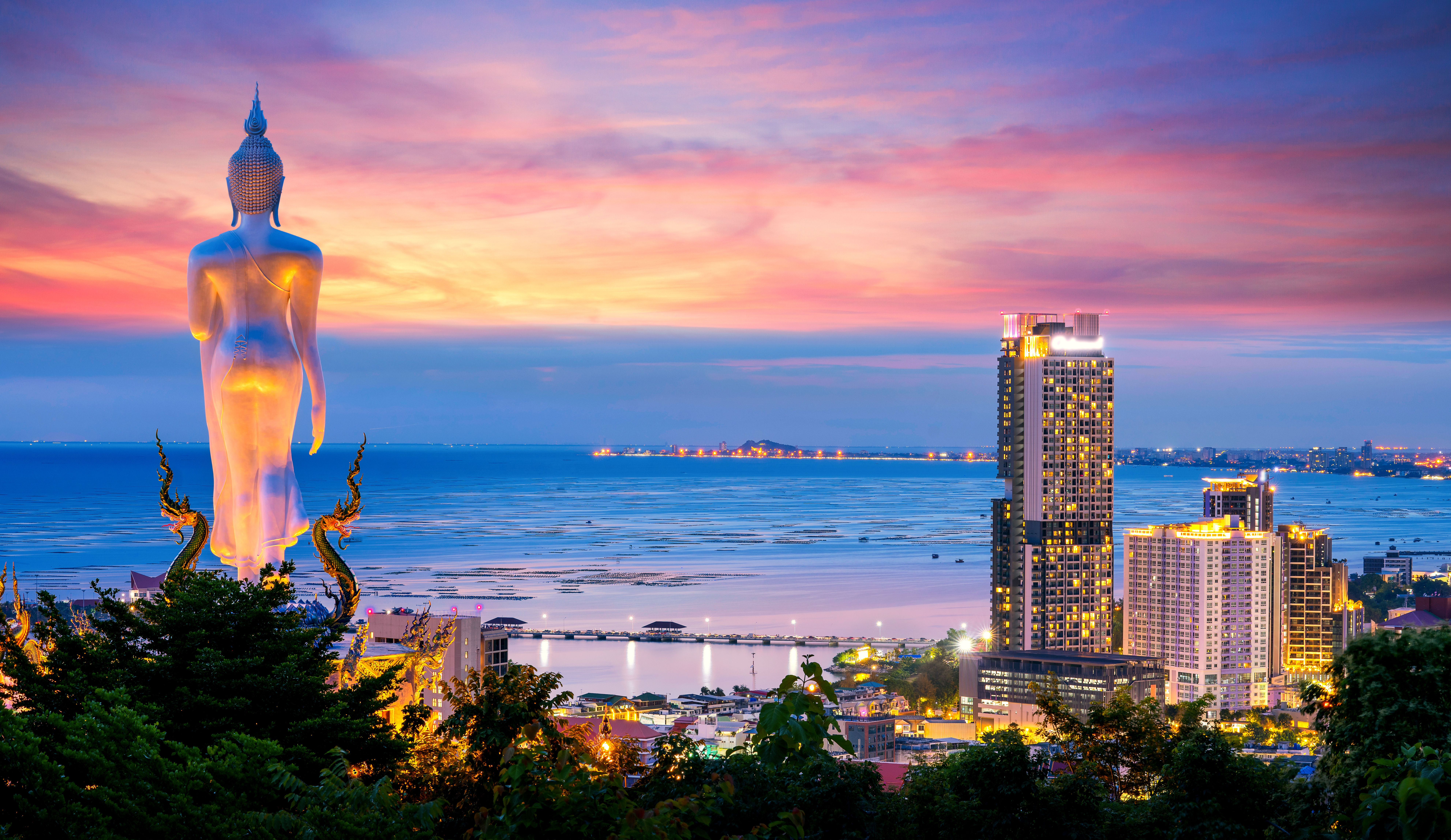 Panoramic view overlooking Pattaya bay at at sunset with purple and pink sky, huge standing big Buddha and hotels.