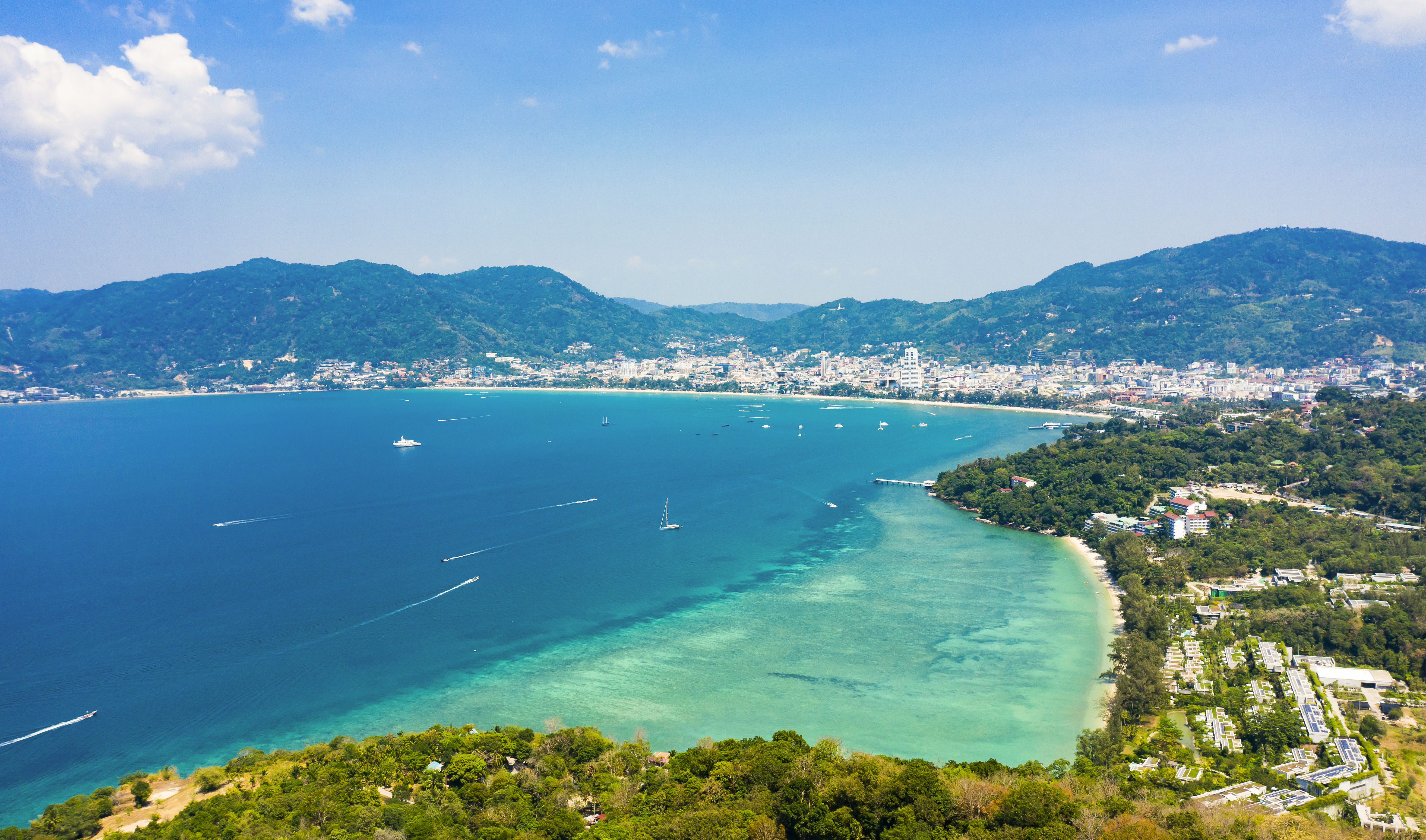 Aerial view of Patong and Tri Trang Beach in Phuket, surrounded by turquoise waters with sail boats, white sand and green hills.