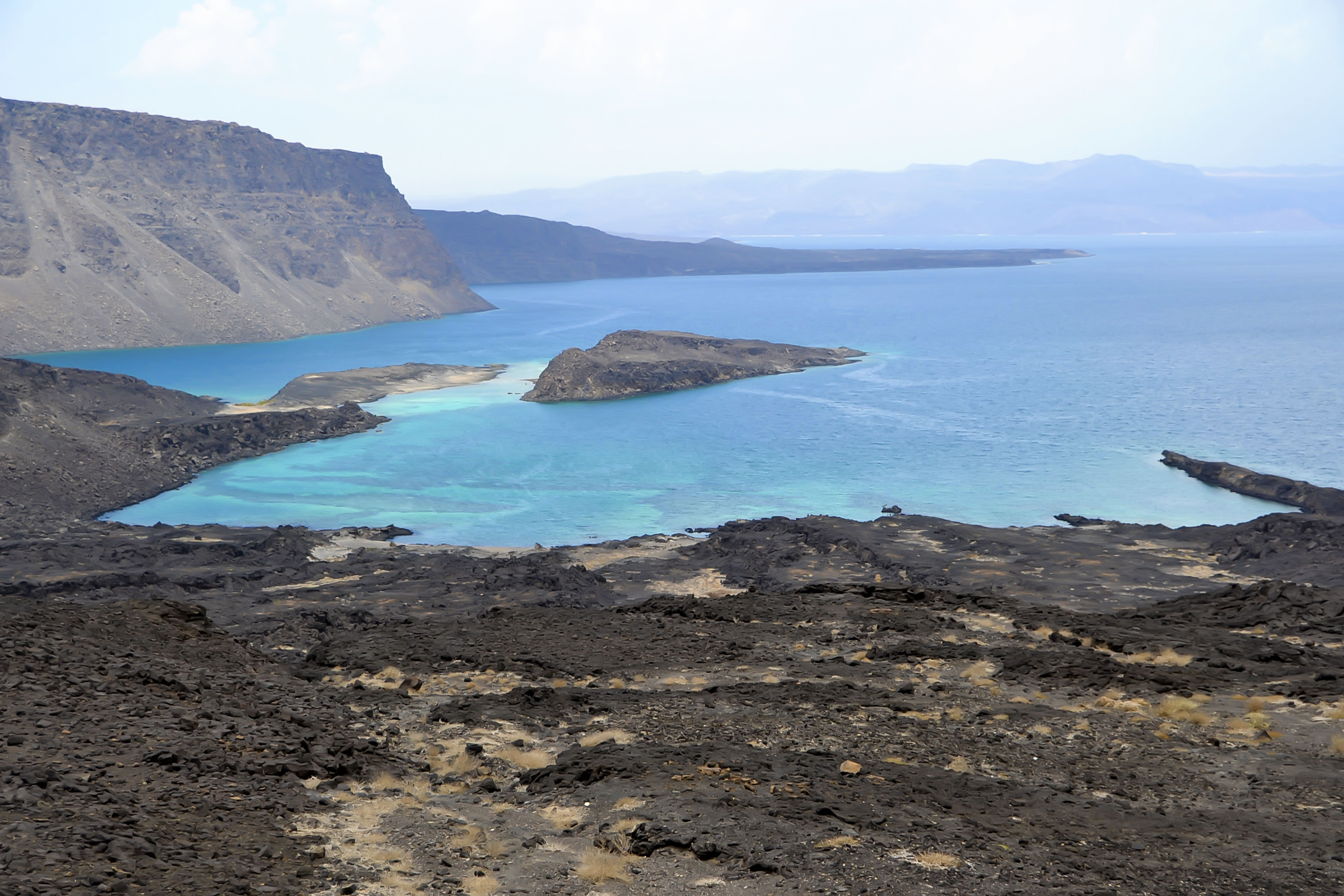 Panorama view of dark sand beach in Djibouti, with blue waters, cliffs and clear skies. 