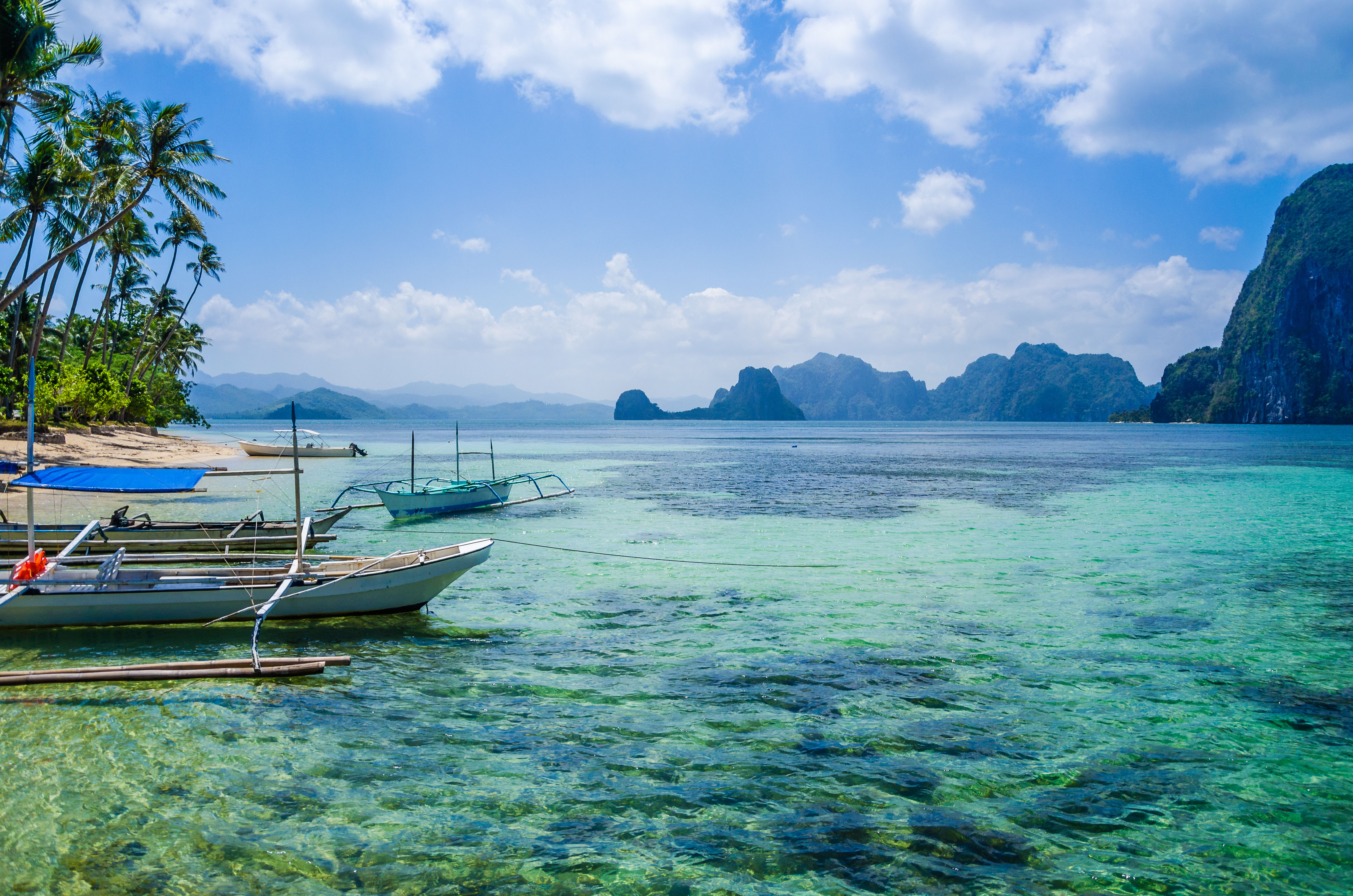Traditional Filipino boats moored on sandy beach with shallow clear turquoise water and limestone cliffs in the background, Coron, Philippines. 