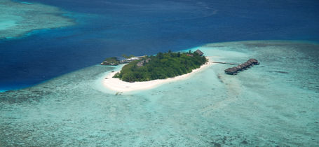 Small remote island with resort, covered in green tropical trees with white sand beaches and surrounded by shallow reefs and turquoise waters in the Maldives.