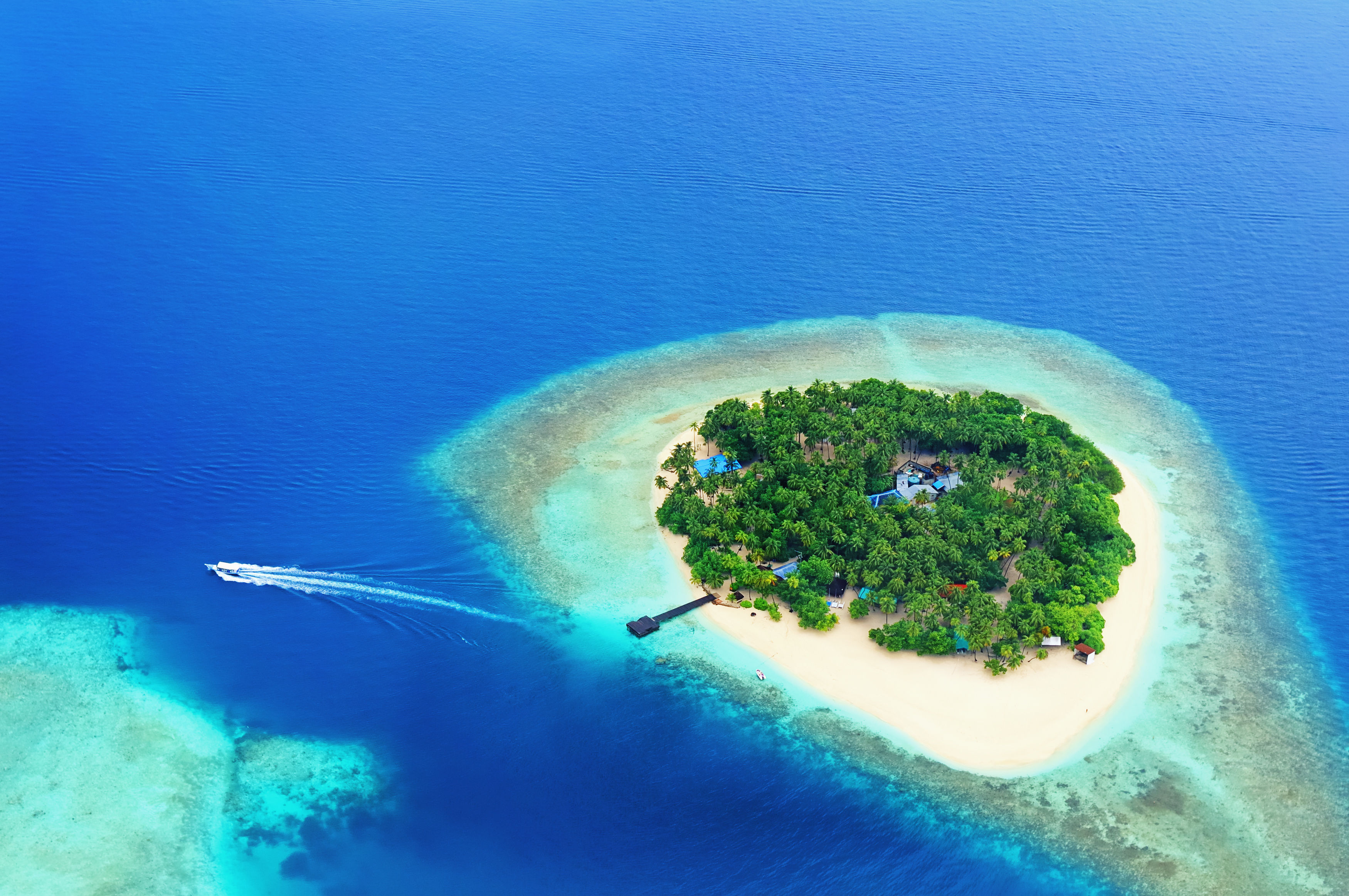 Aerial view of beautiful Maldives island covered in green trees and surrounded by white sand beaches, turquoise waters and with pier and speed boat. 