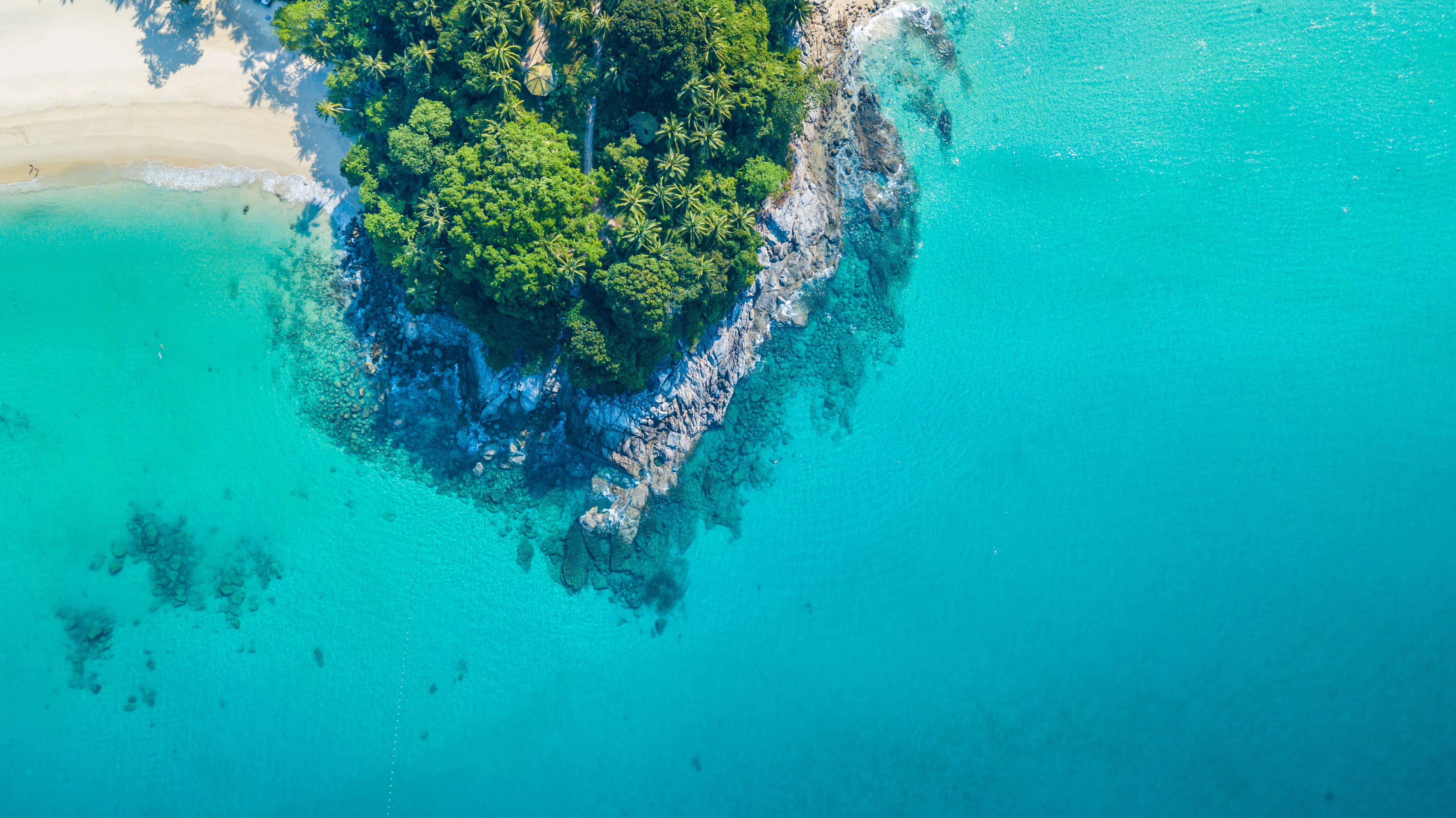 Aerial drone view of rocky outcrop with white sand beach and turquoise clear water with shall reef in Phuket, Thailand.
