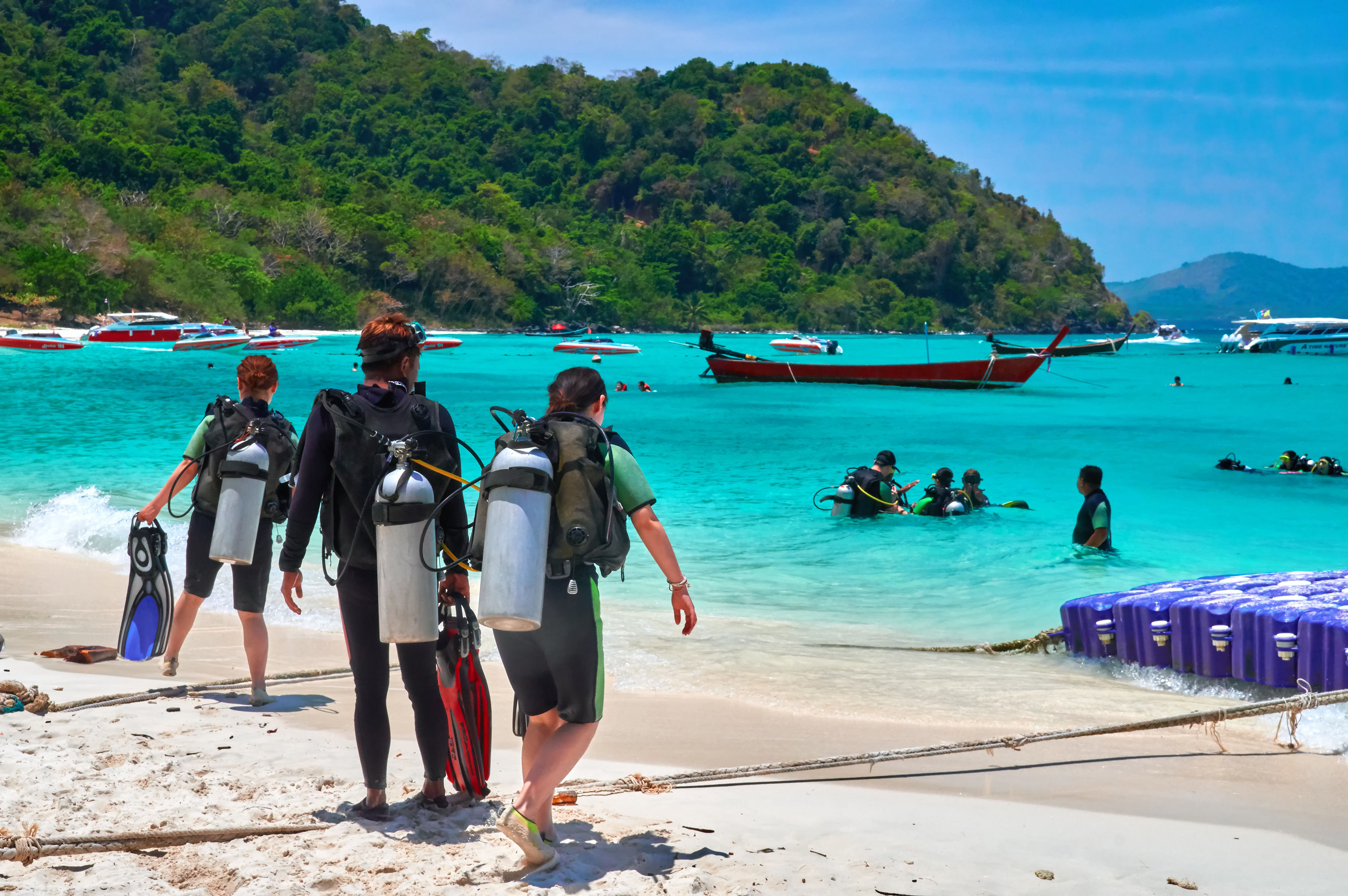 Scuba divers on white sand beach walking towards turquoise bay with divers learning to dive in shallow water.